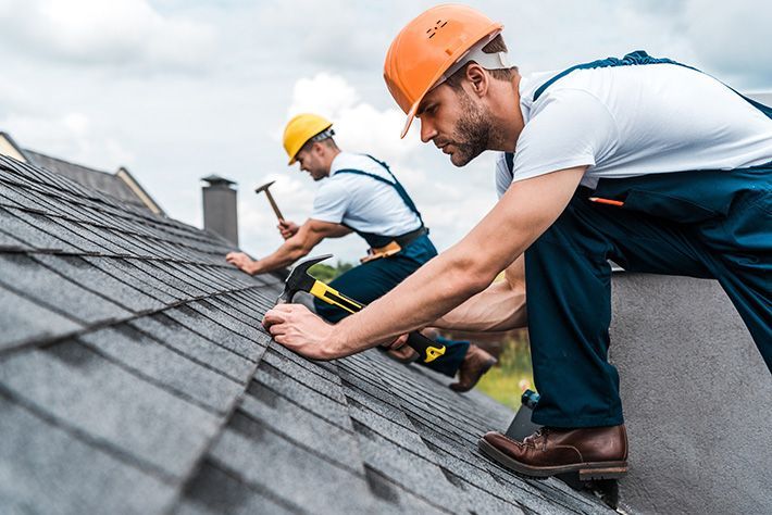 Worker in a neon safety vest repairing a shingled roof with a power tool.