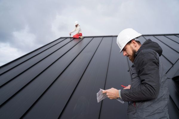 Worker in a hard hat inspecting a dark metal roof with a clipboard under cloudy skies