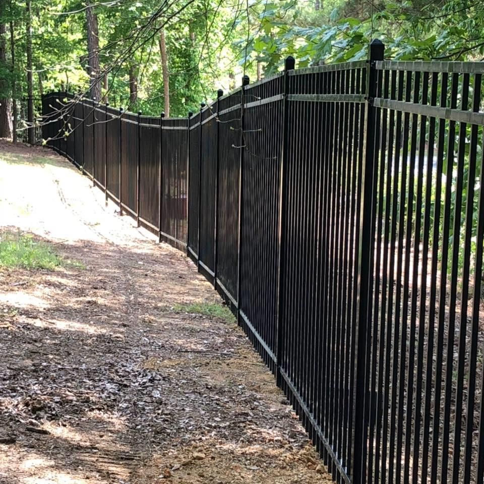 Black metal fence curves along a dirt path in a wooded area.