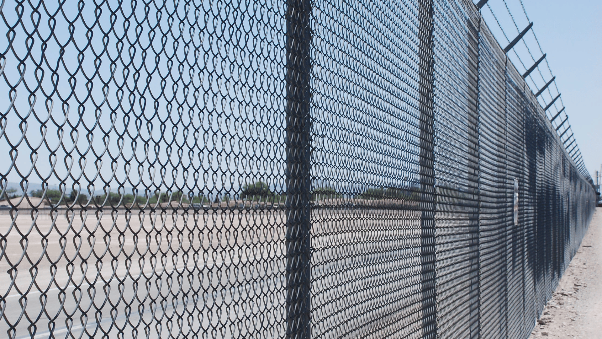 Chain-link fence with barbed wire on top, enclosing a barren landscape.