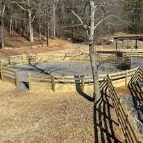 Wooden fenced riding arena in a wooded area with trees and sunlight.