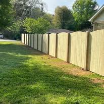 A wooden privacy fence in a yard with green grass, trees, and a house in the background. Sunny day.