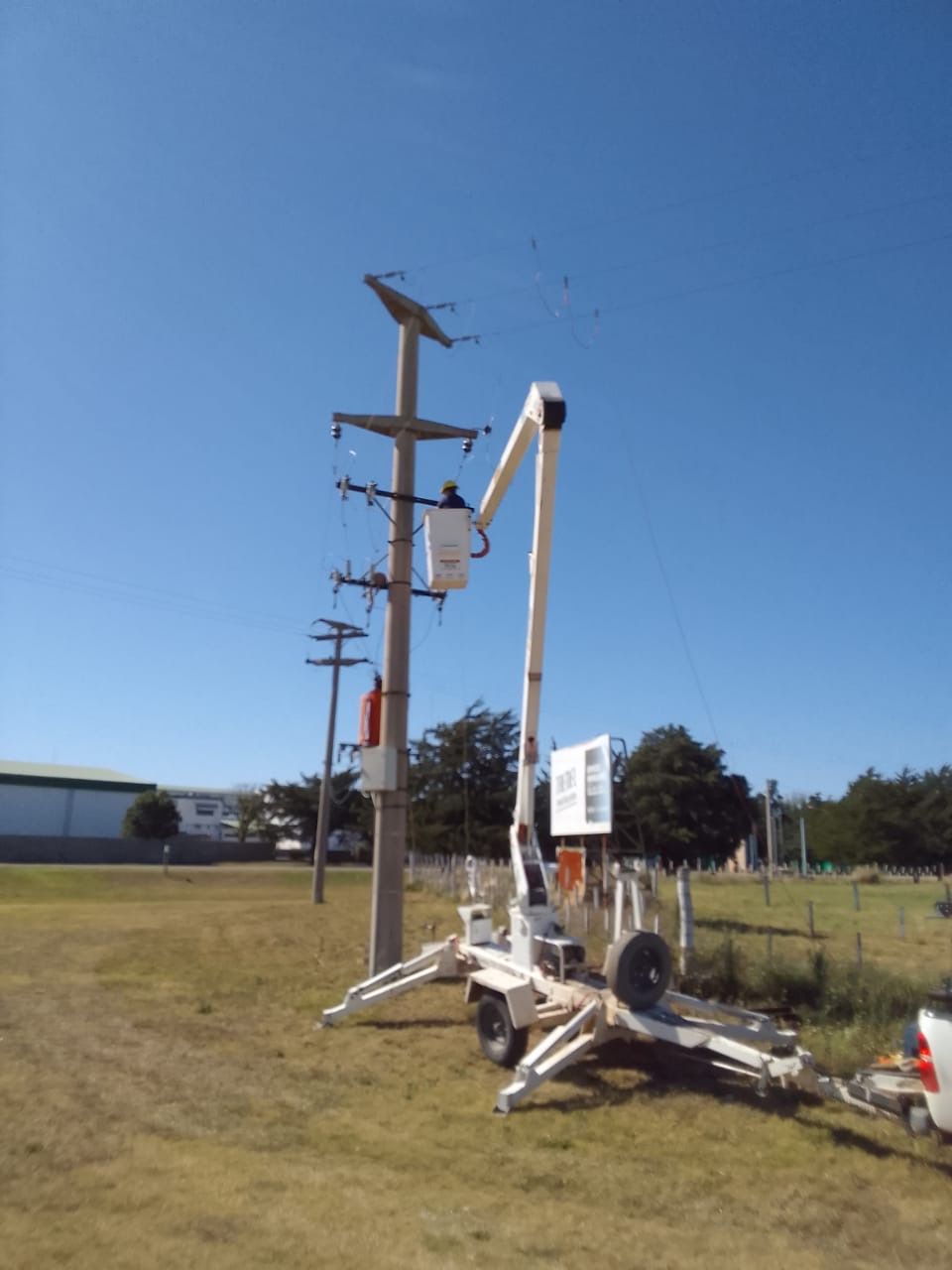 Camión utilitario con pluma elevada que presta servicio a líneas eléctricas en un poste de madera bajo un cielo azul.