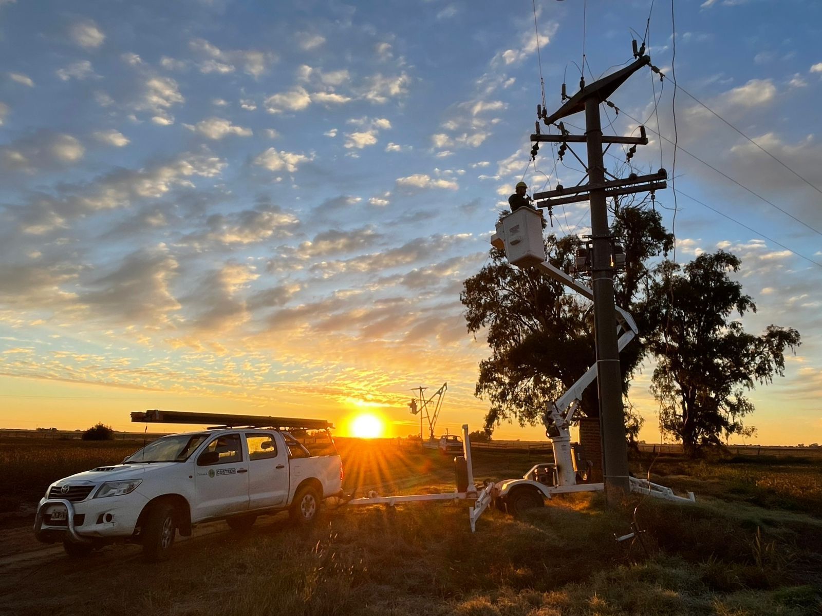Electricista trabajando en un poste de electricidad al amanecer con un camión de servicios públicos en un entorno rural.