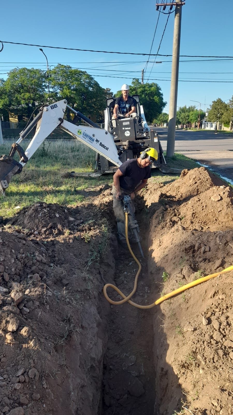 Dos trabajadores excavando una zanja cerca de líneas eléctricas, utilizando una pequeña excavadora