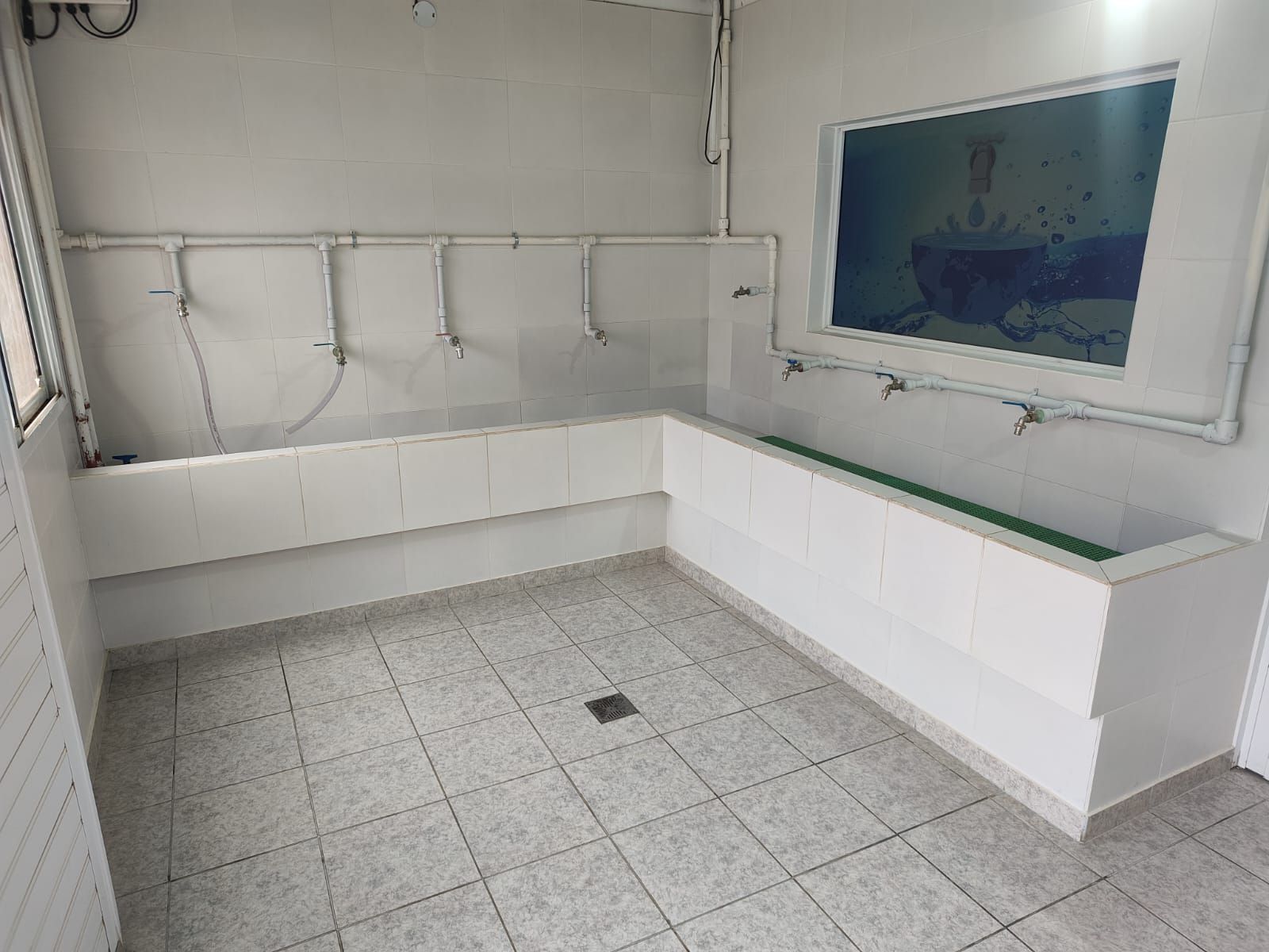 Habitación con azulejos blancos, varios lavabos, ventana y desagüe.