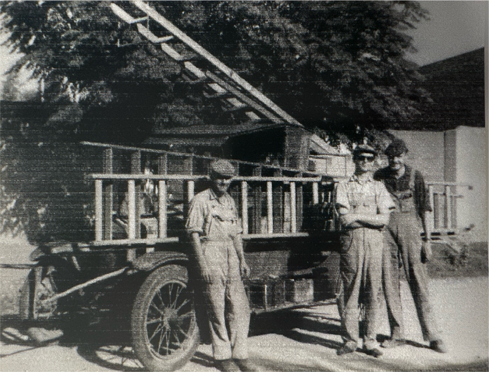 Tres bomberos posan con un camión de bomberos antiguo; una escalera alta está montada en el camión.