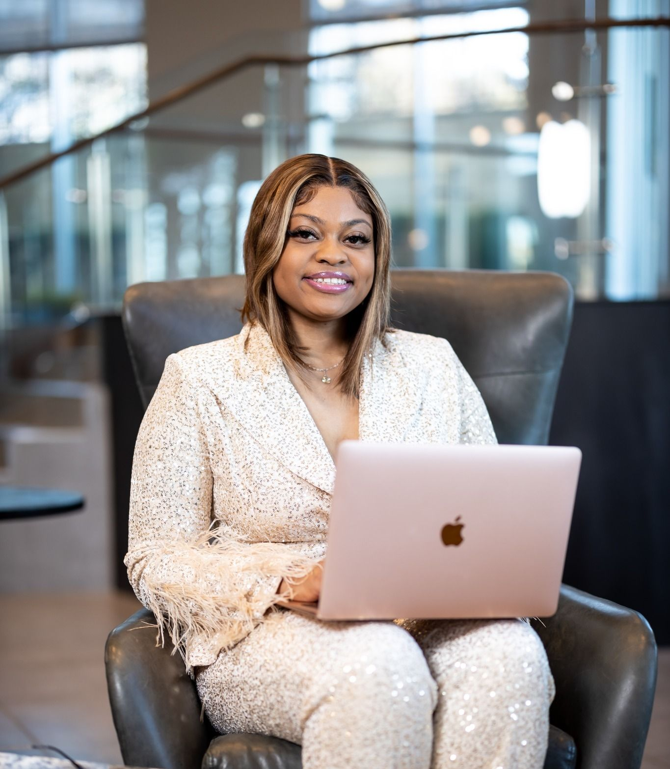 Woman in a sparkling cream suit sits in a chair, holding a laptop. She smiles in a modern building.