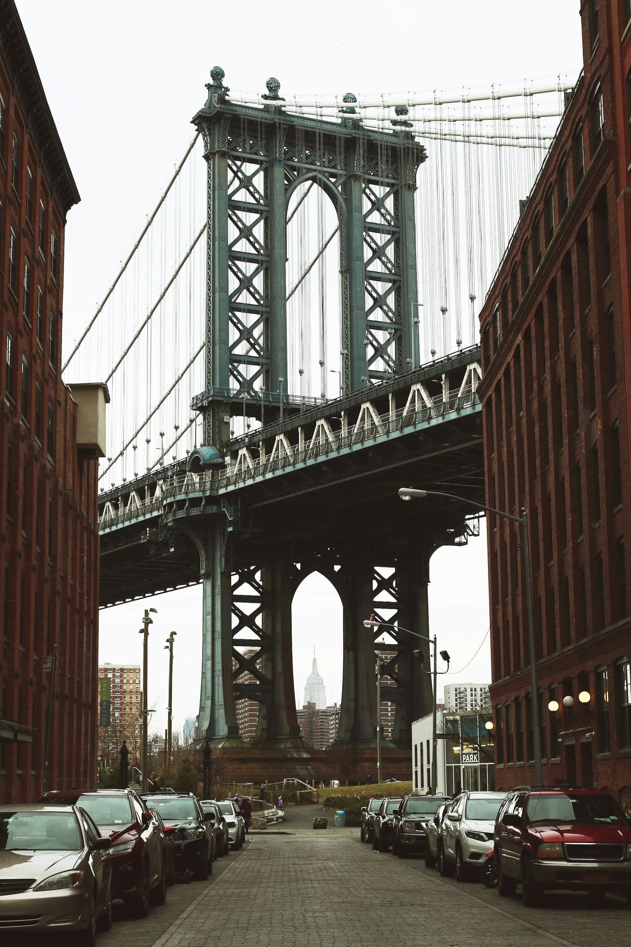 Manhattan Bridge viewed from a street lined with parked cars and buildings.