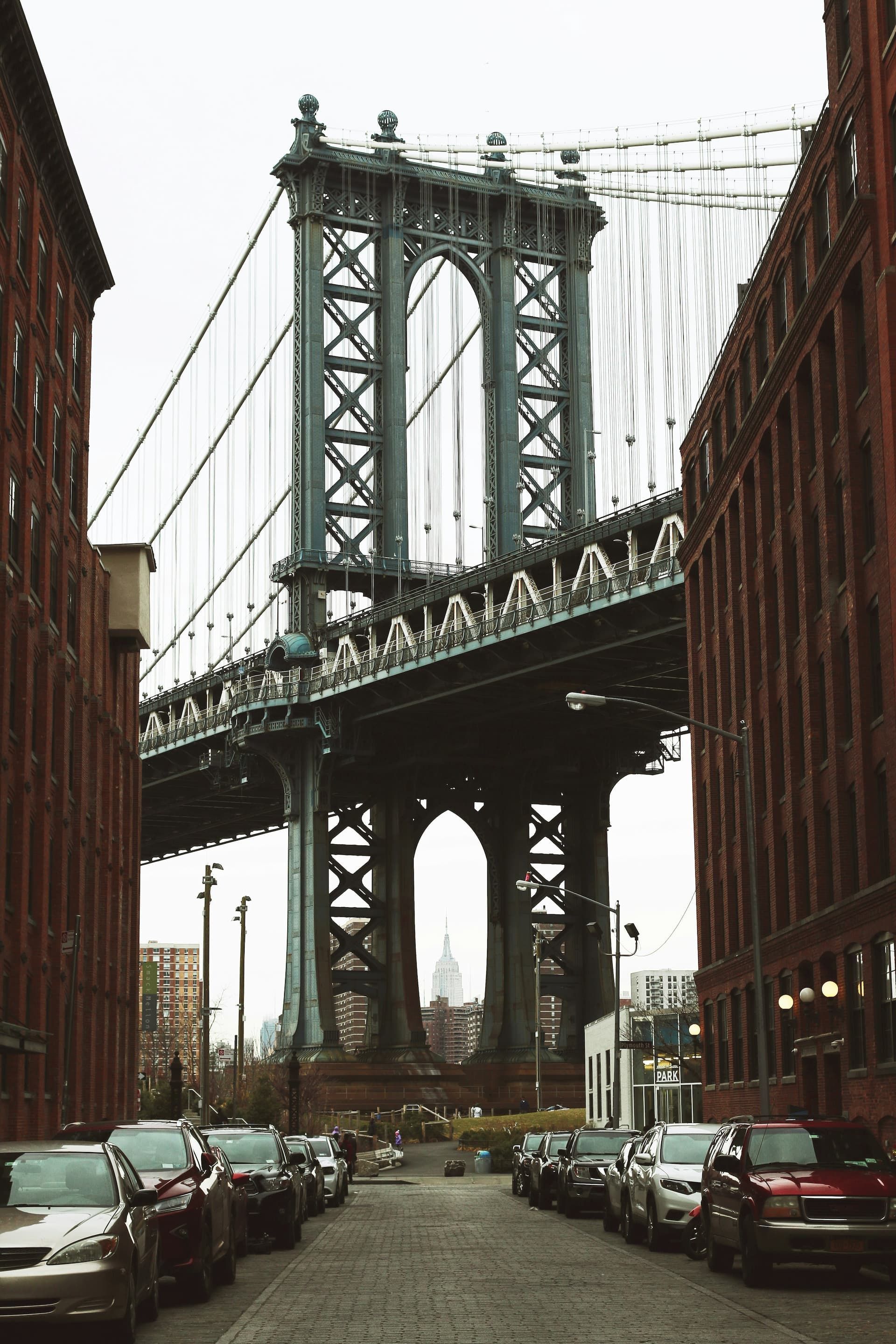 Manhattan Bridge viewed from a street lined with parked cars and buildings.