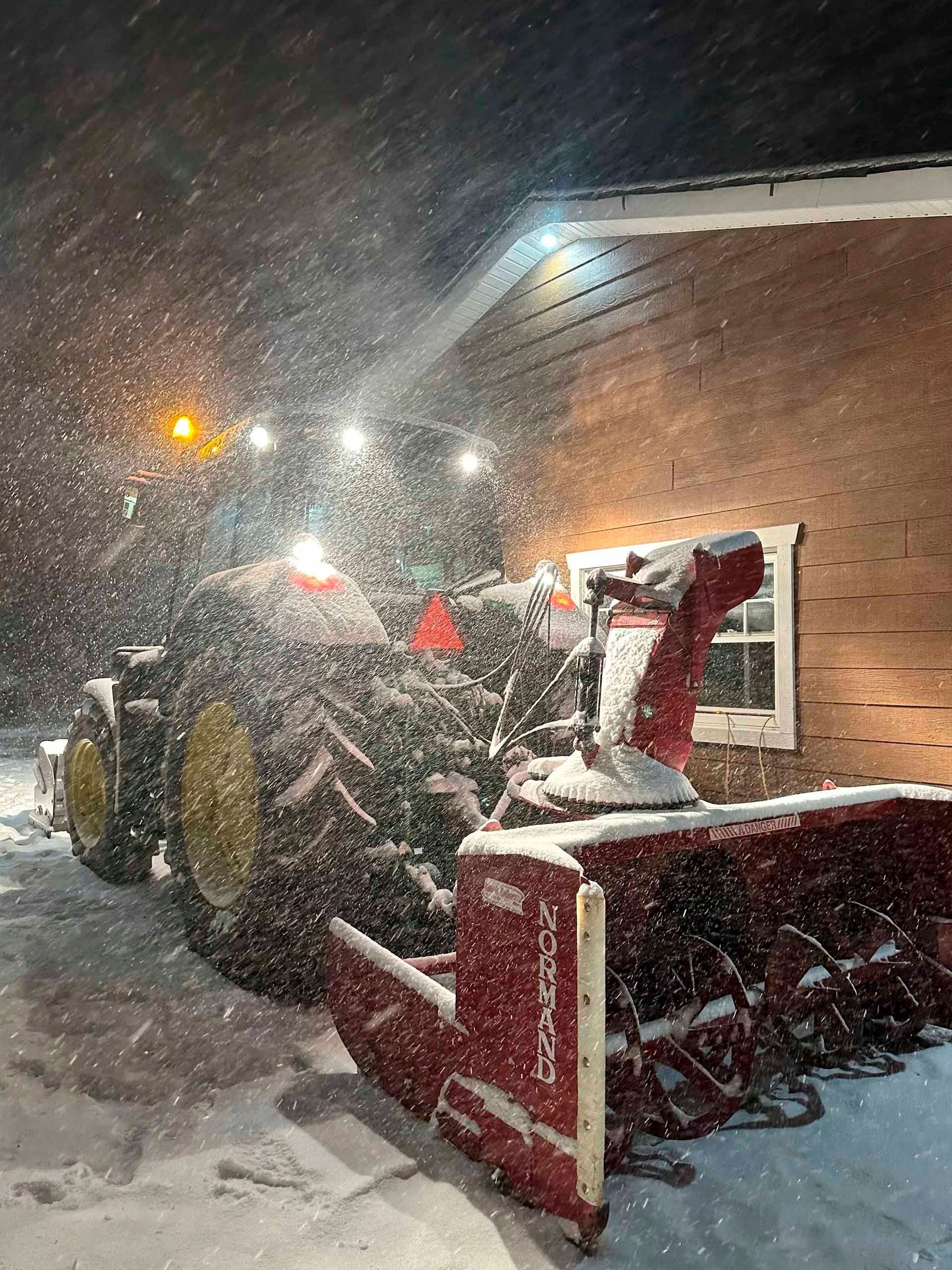 Un tracteur équipé d'une souffleuse à neige dégage la neige près d'un bâtiment, de nuit, éclairé par des projecteurs. Il neige abondamment.