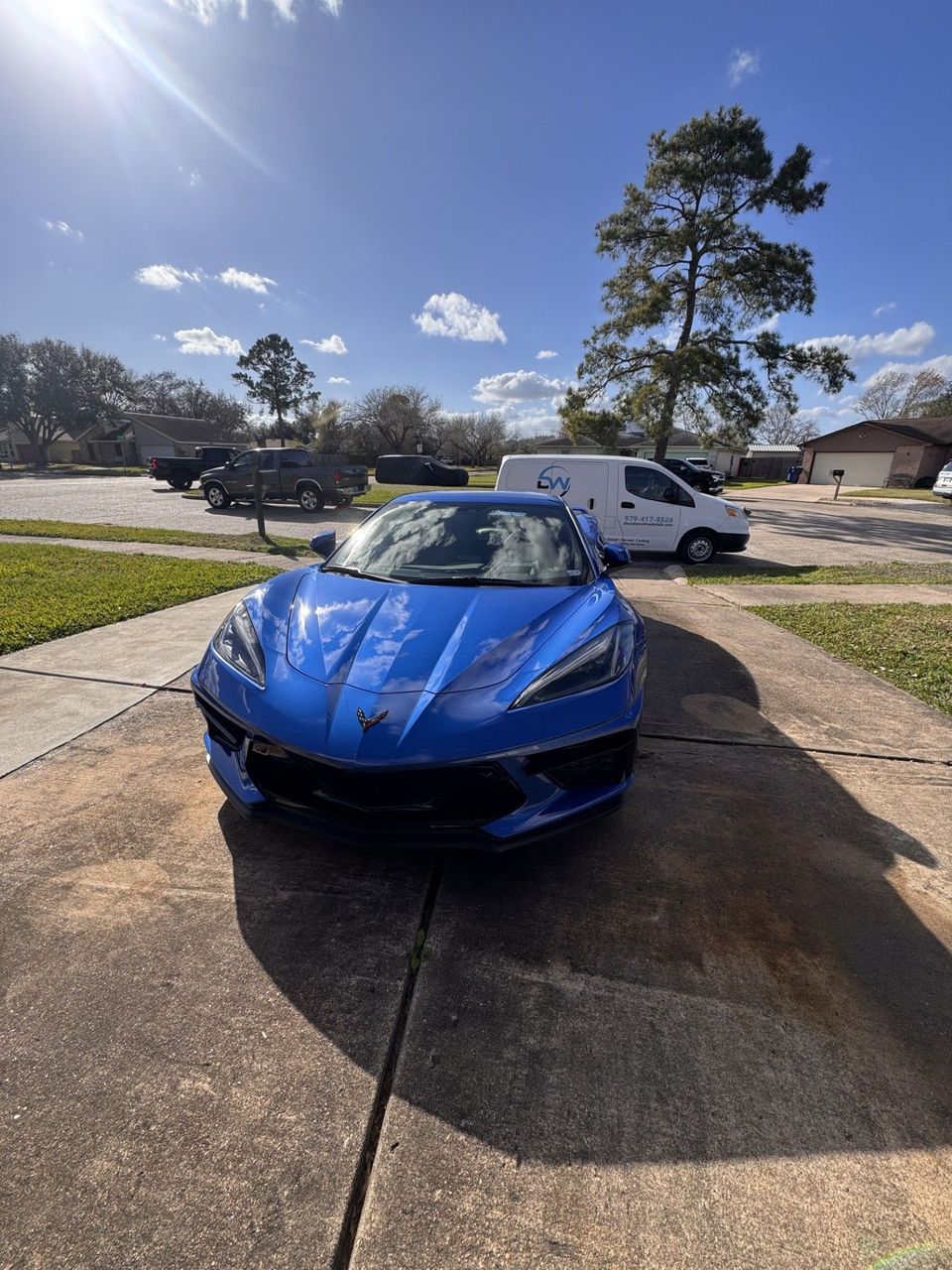 Blue sports car parked in a driveway on a sunny suburban street, with a white van nearby.