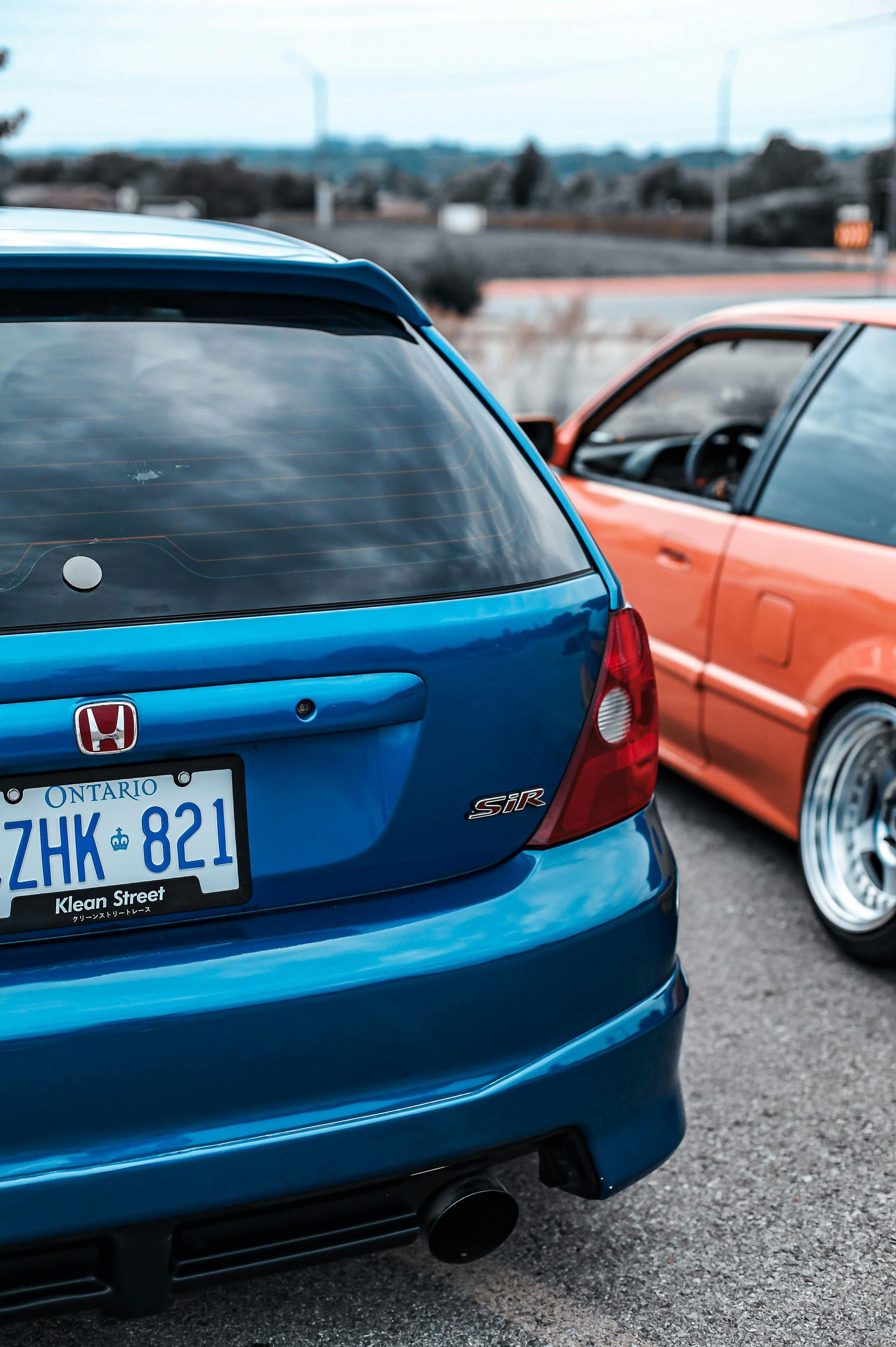 Blue Honda hatchback and orange car parked side by side in an outdoor lot