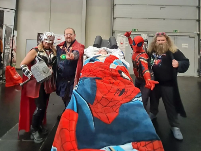 Eine Gruppe von Menschen in Superheldenkostümen, die um einen Schützling im Transportsessel stehen.