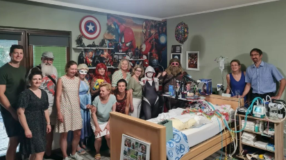 Eine Gruppe von Menschen, teils kostümieert, steht um ein Krankenbett herum.