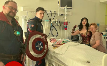 Eine Gruppe von Menschen in Superheldenkostümen steht um ein Kind in einem Krankenhausbett herum.