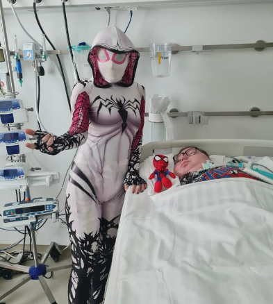 Eine Frau in einem Spider-Man-Kostüm steht neben einem Kind in einem Krankenhausbett.