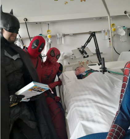 Eine Gruppe von Menschen in Superheldenkostümen steht um ein Krankenhausbett herum.