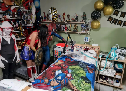 Eine Gruppe von Menschen in Superheldenkostümen steht um ein Krankenbett herum.