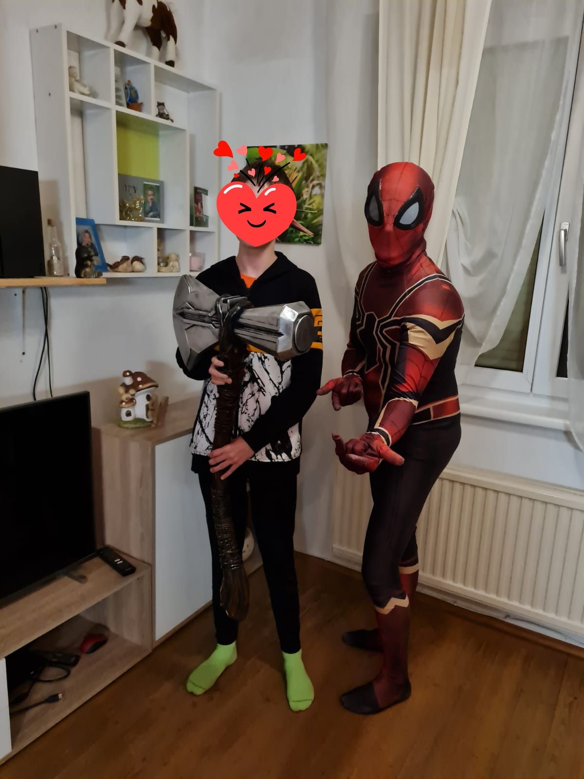 Ein Man und ein Schützling in Superheldenkostümen stehen nebeneinander in einem Wohnzimmer.