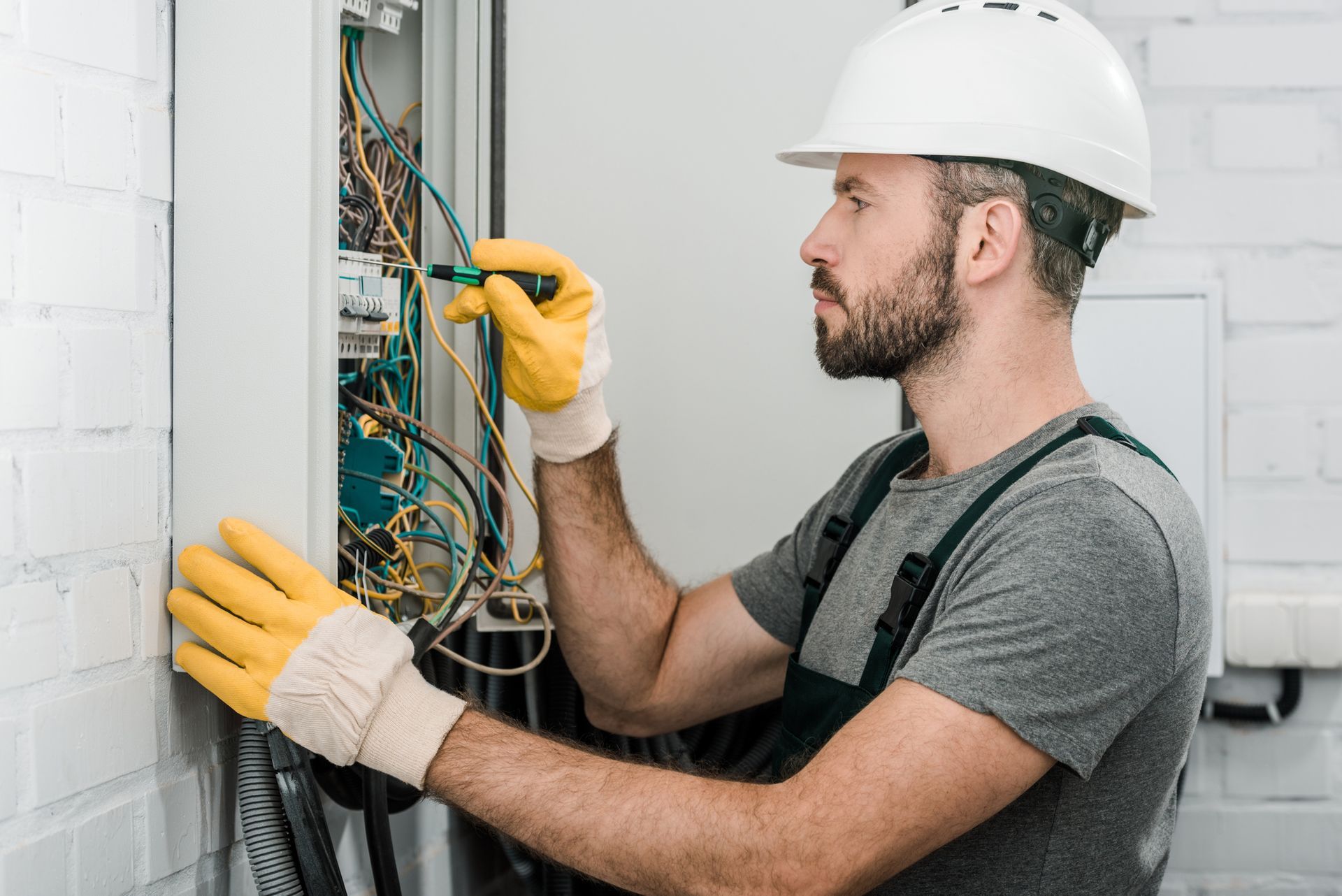Electrician service technician wearing gloves and helmet working on a fuse box wiring panel. Electrician service technician wearing gloves and helmet working on a fuse box wiring panel.