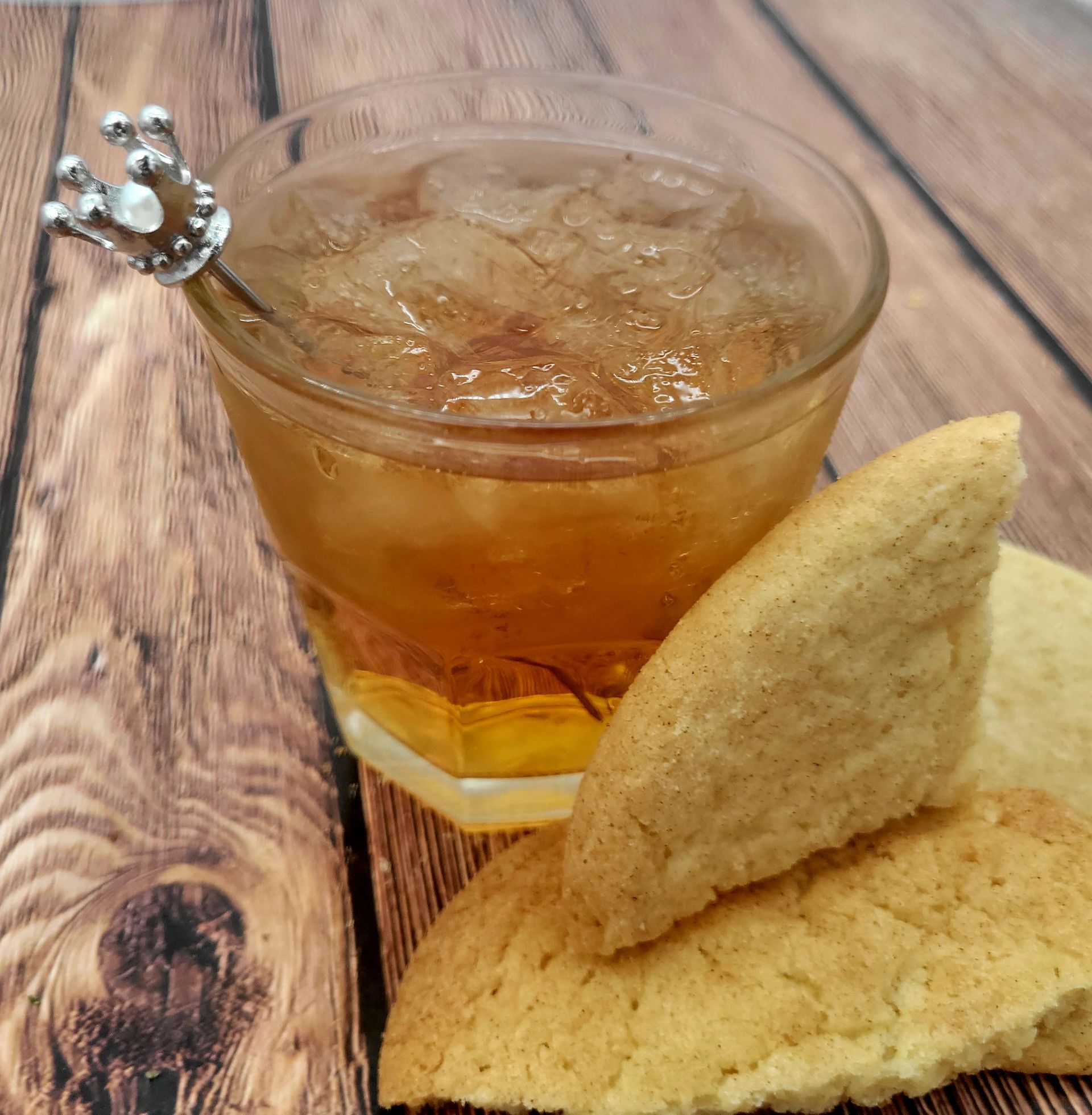 A glass of alcohol with a silver crown and cookies on wooden table.
