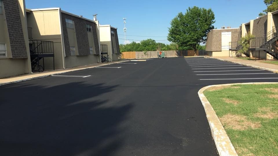 Apartment complex parking lot with fresh black asphalt, beige buildings, and a tree-lined edge under clear daylight