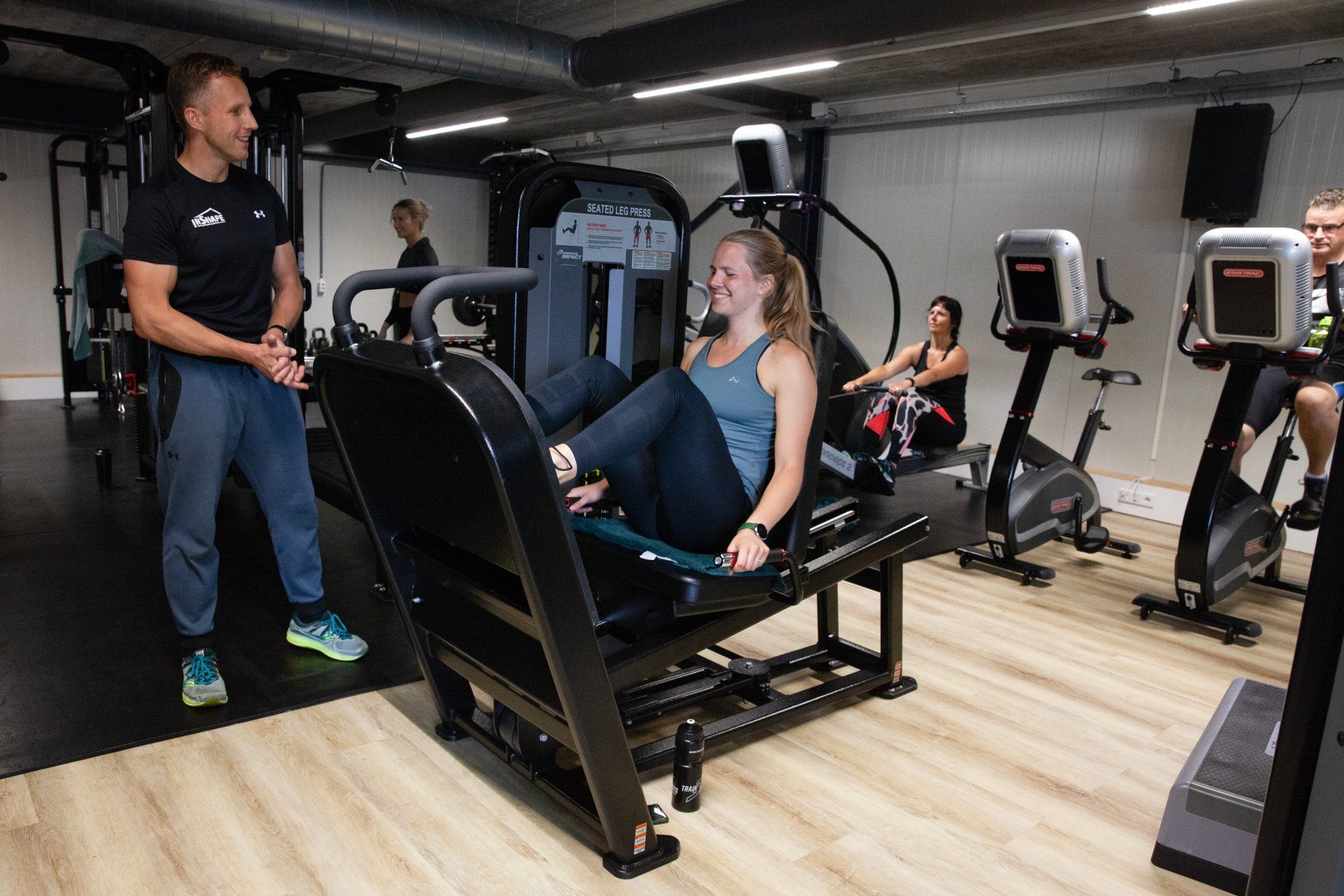 Een vrouw zit op een fitnessapparaat in een sportschool terwijl een man achter haar staat.