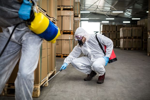 Two people in hazmat suits spraying insecticide in a warehouse with stacked boxes.