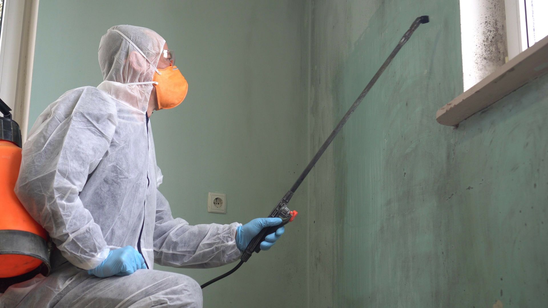 Person in protective suit spraying a wall with a long nozzle. Orange tank and mask, teal wall, window.