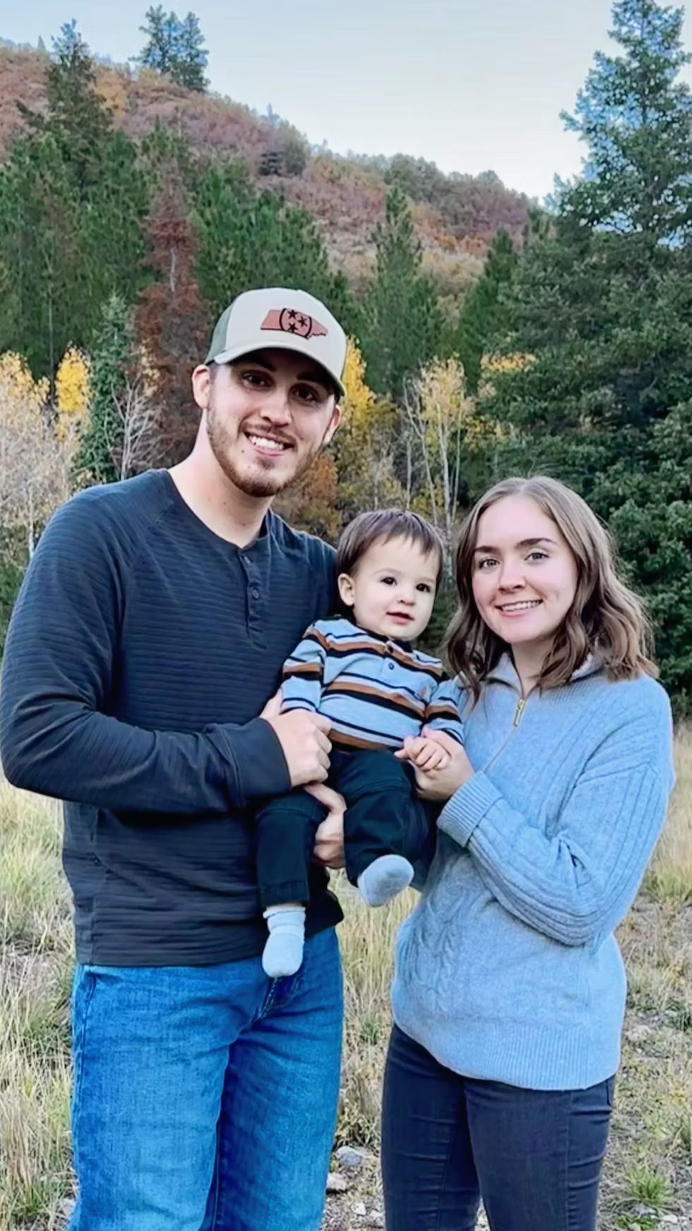 Family of three smiles for the camera. The man holds the child. Autumn trees in the background.