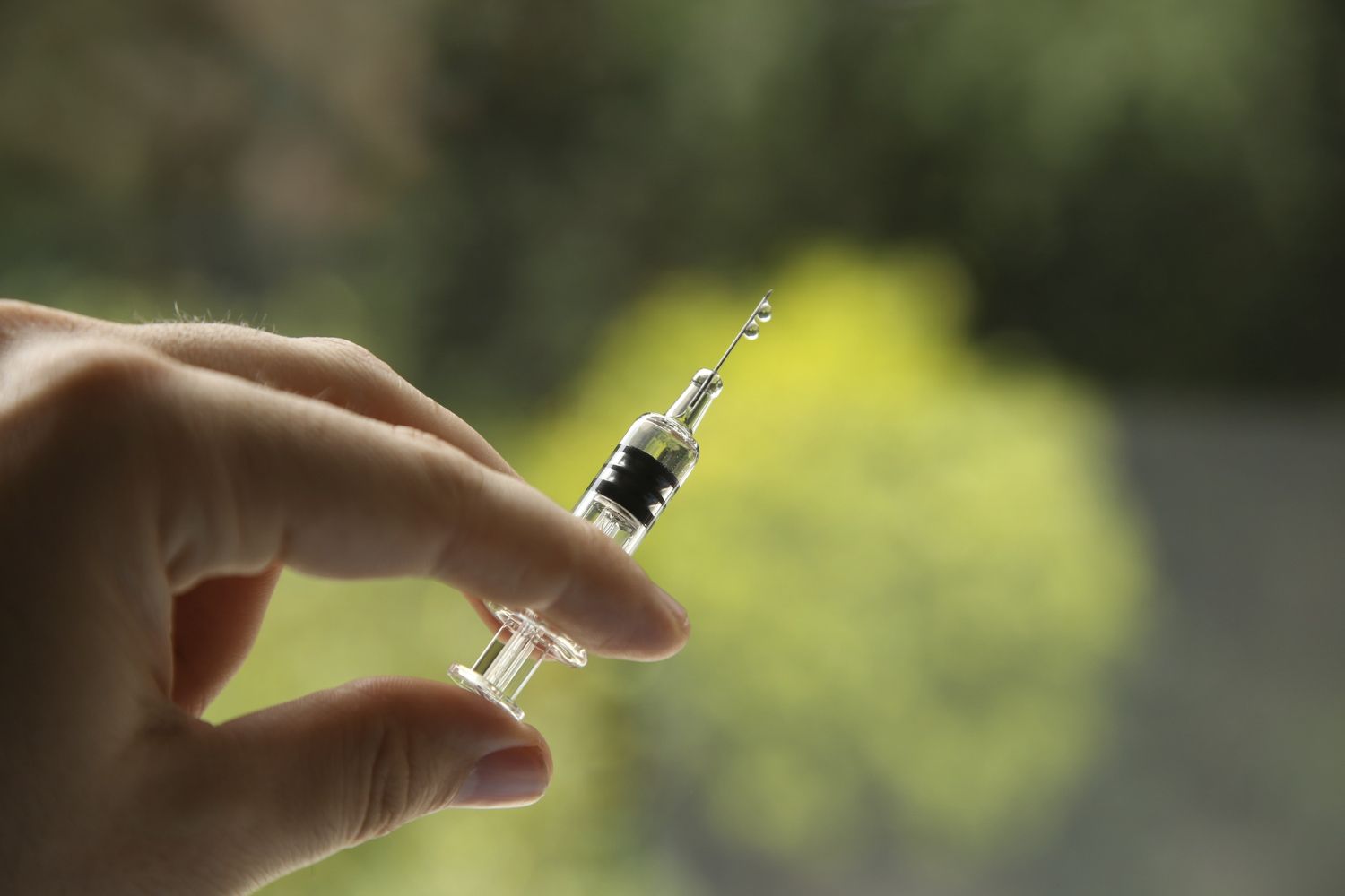 Hand holding a syringe, a drop of liquid at the needle's tip, against a blurred green background.