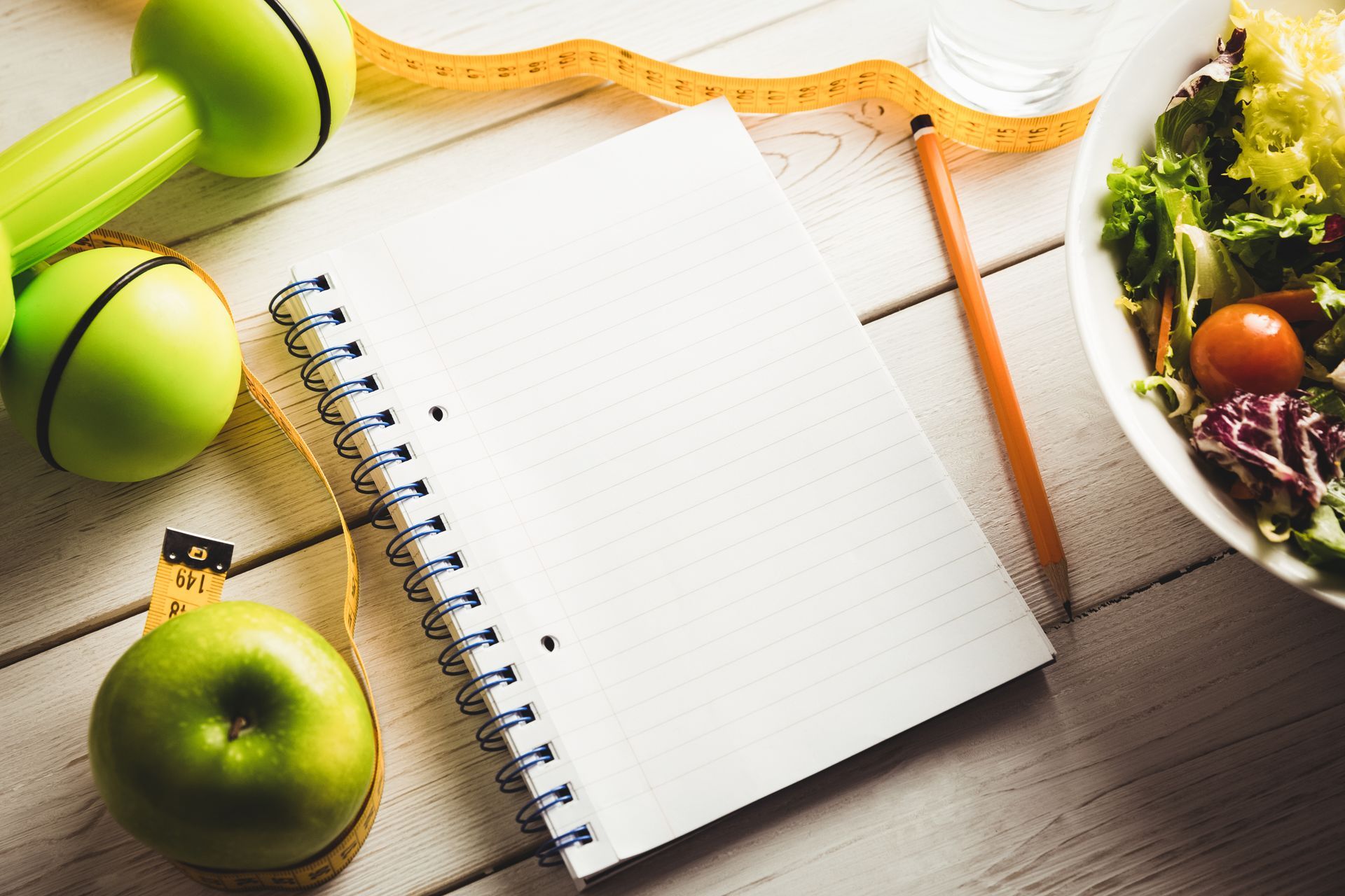 A notebook, measuring tape , dumbbells, an apple and a bowl of salad are on a wooden table.