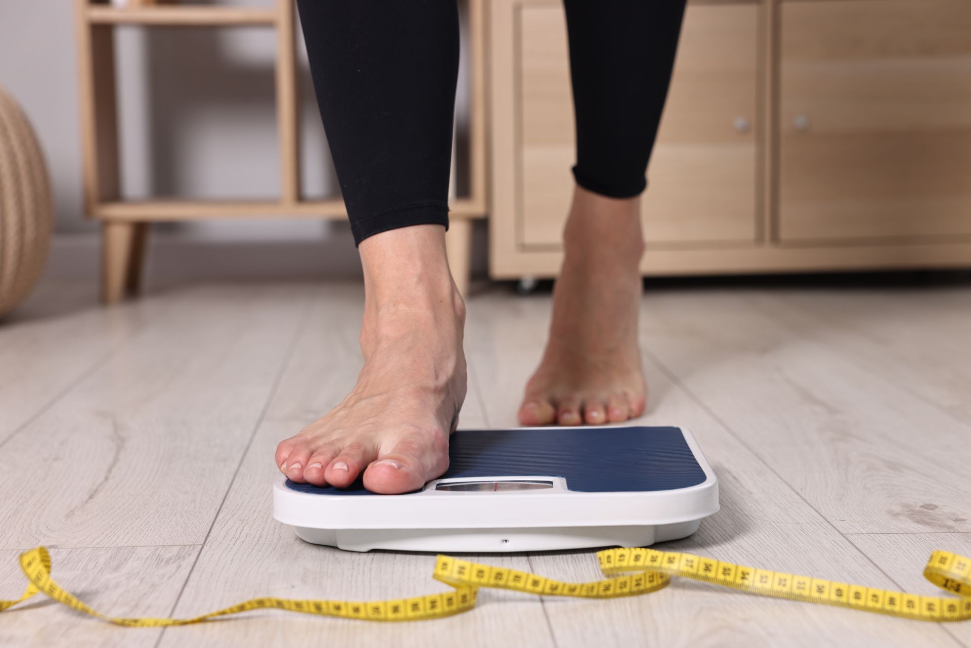 Person stepping onto a bathroom scale. Legs are in black leggings; a yellow tape measure is on the floor.