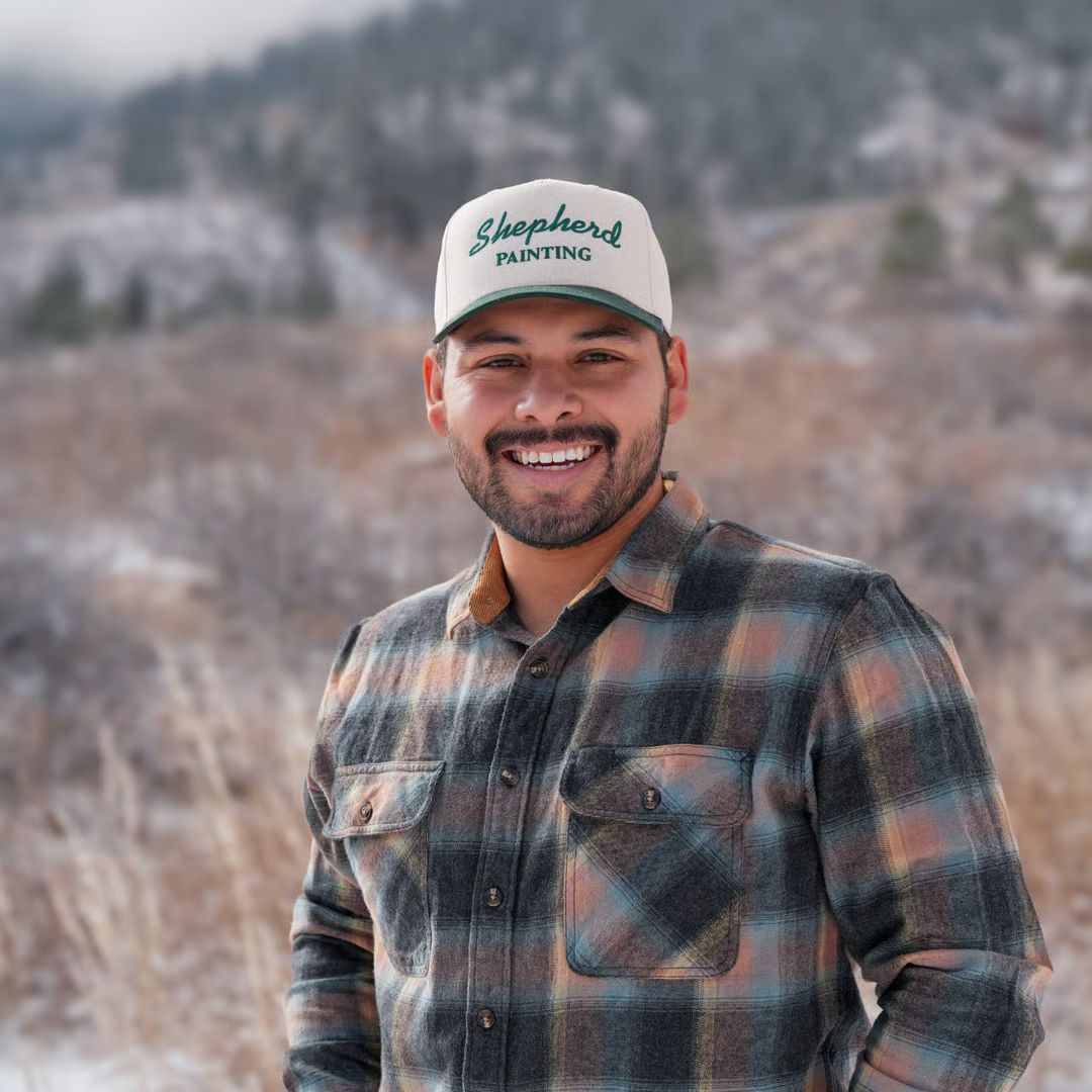 Smiling man in a plaid shirt and cap standing outdoors in a snowy mountain setting