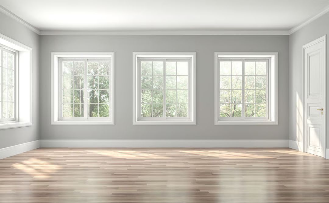 Empty bright room with gray walls, white trim, large windows, and light wood floors.