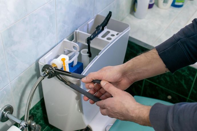 Hands using pliers to tighten a metal hose connection on the side of an open toilet tank in a tiled bathroom.