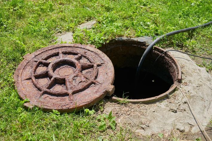 An open, rusted circular metal manhole cover sits on the ground next to an open, dark maintenance hole with a hose inside.