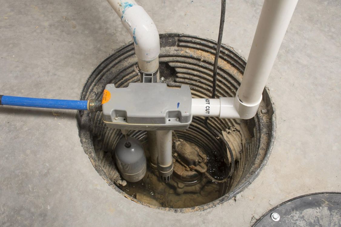 A sump pump installed in a circular concrete pit, featuring a gray pump housing, white PVC pipes, and a blue water hose.