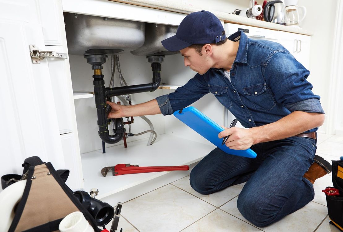 A professional plumber in work clothes kneeling under a kitchen sink, examining the pipes while holding a blue clipboard.