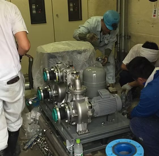 Workers in a technical facility install a metallic pump system mounted on a gray frame.
