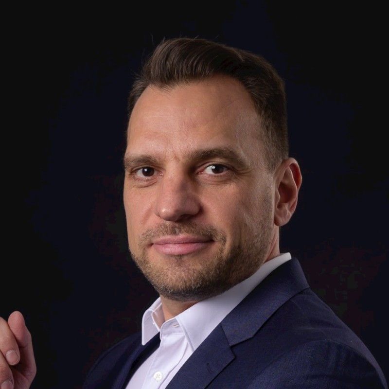Man in a suit smiles, headshot. Dark background, white shirt, dark blue jacket.