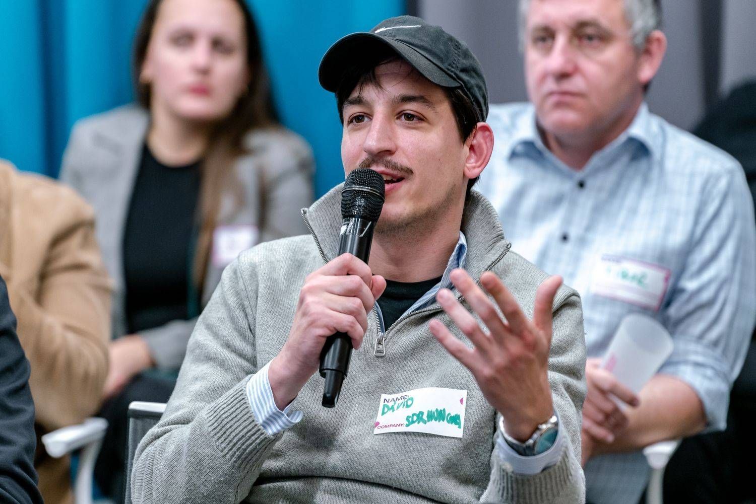 Man with microphone, wearing hat and sweater, speaking at event. Others seated nearby.