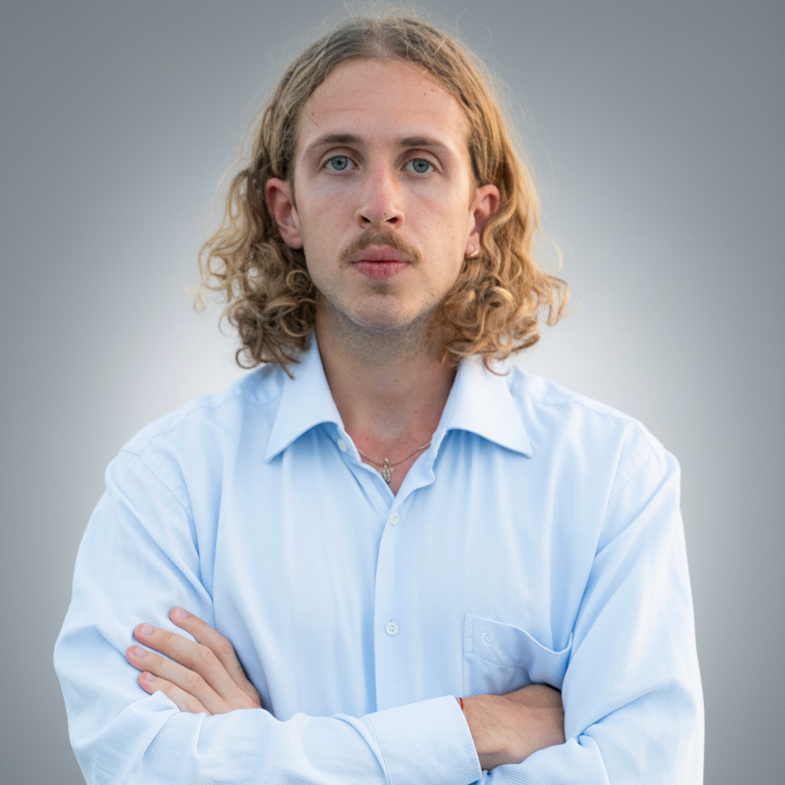 Man with curly blonde hair and a mustache wearing a light blue button-up shirt, arms crossed.