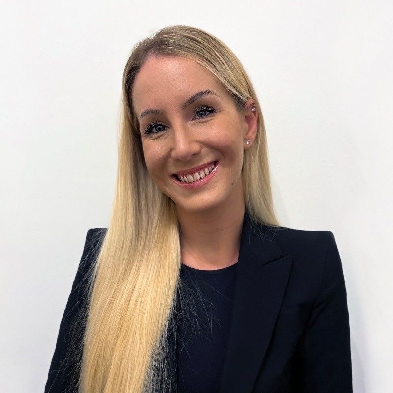Blonde woman smiles at camera, wearing a navy blazer and top, against a white background.