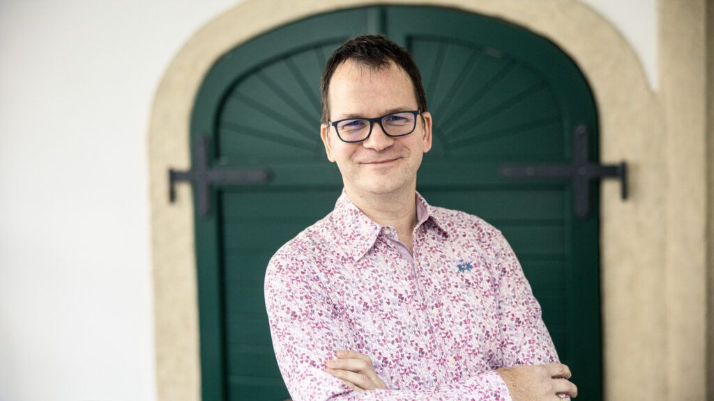 Man in glasses, pink patterned shirt, arms crossed, smiling. Leaning near a green door.