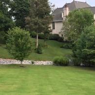 A large house with a grey roof sits in the background of a lush green lawn, separated by a stone-lined drainage bed.