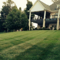 A two-story house with a large balcony and covered patio overlooks a wide, grassy hill with visible lawnmower stripes.