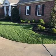 A red brick house with a green lawn, dark mulch garden beds, and manicured shrubs under a bright sky.
