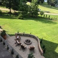A curved stone patio with a fire pit and four wooden chairs overlooking a large, grassy lawn with trees in the distance.